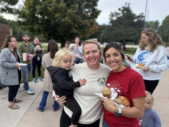 Group of people outdoors; two women smiling and holding food. A child is in the arms of one woman. Background includes others standing, talking, and carrying items.