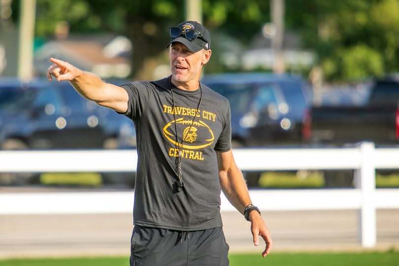 A man in a black hat and shirt stands on a field, pointing. A white fence is in the background.