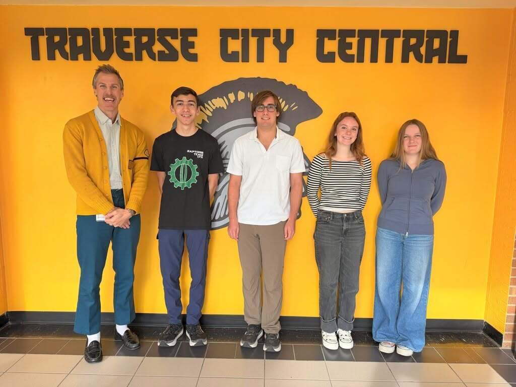Five people stand in front of a yellow wall reading "Traverse City Central". They wear various clothes and shoes, standing in a line.