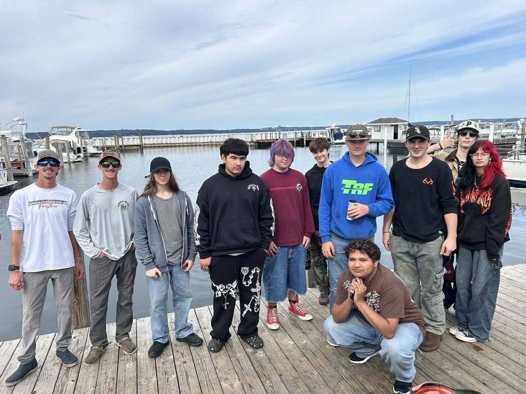 Group of individuals, some in hats, posing on a wooden dock over water, with boats in the background.