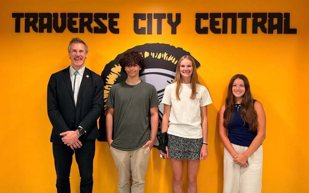 Four individuals stand in front of a yellow wall with "TRAVERSE CITY CENTRAL" in bold letters.