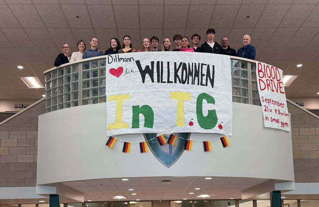Group of people on a balcony with banners, one reading "Willkommen" and another "In T.C." Bright lighting.