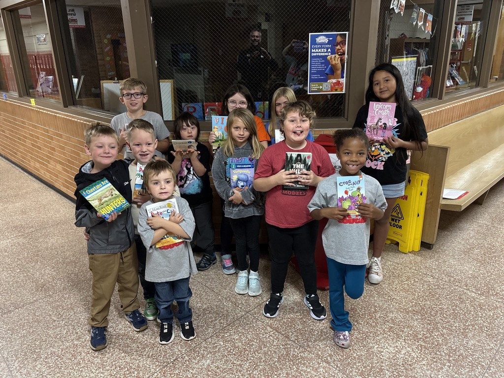 A group of children holding books stands in front of a building with brick walls. Behind them, a person is near a glass window.
