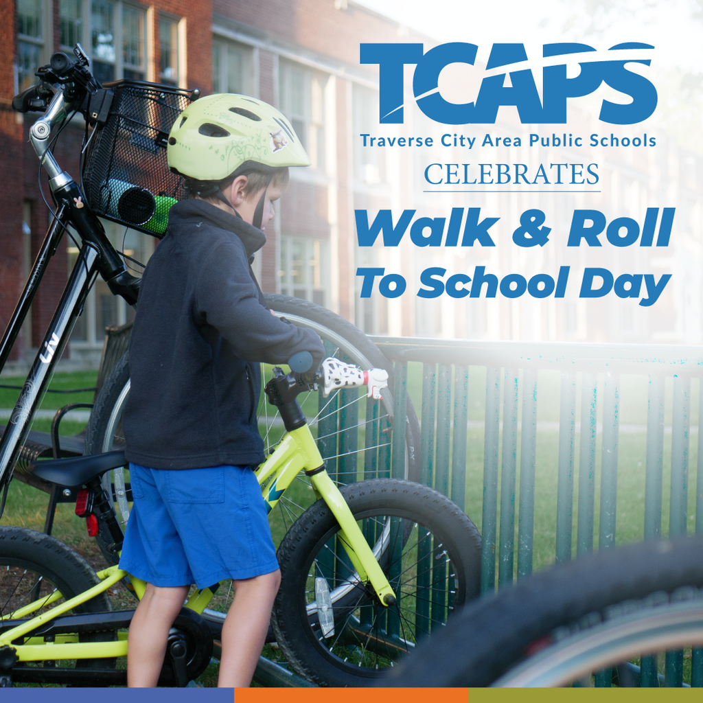 A boy in a helmet holding a yellow bike stands near a school fence with a sign reading "Walk & Roll To School Day."