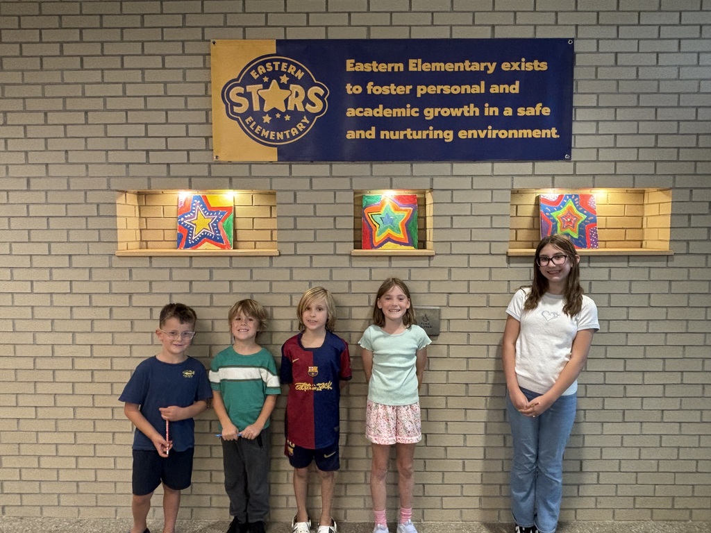 Five children stand in front of a brick wall with a sign reading "Eastern Elementary exists to foster personal and academic growth in a safe and nurturing environment."