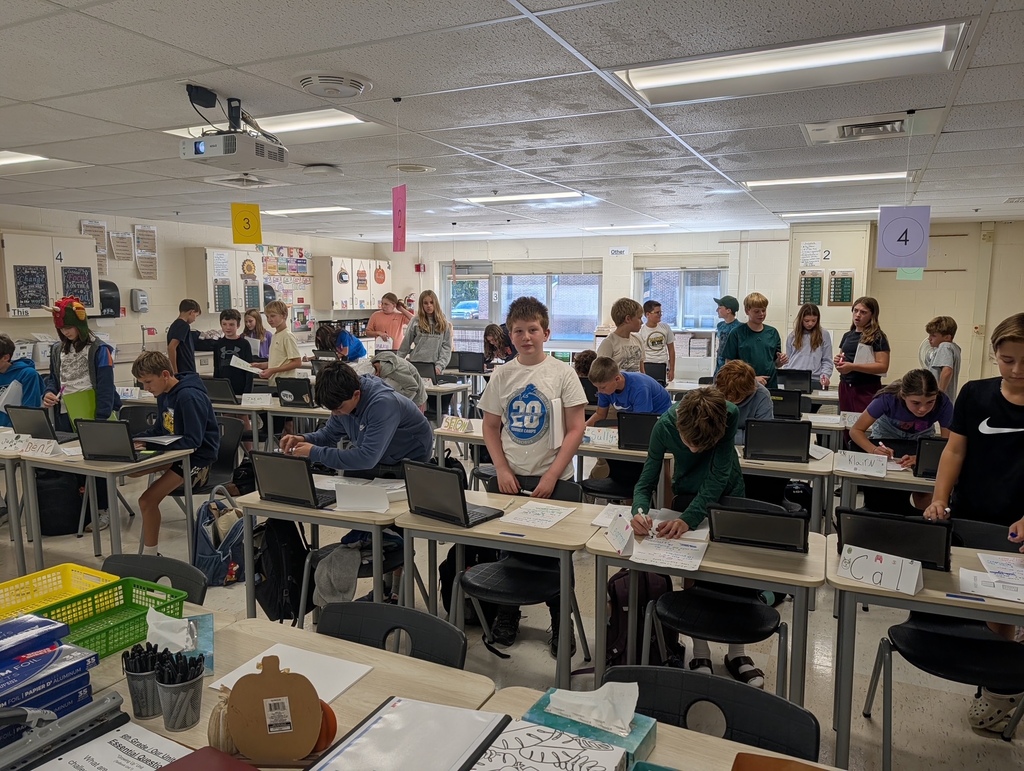 A classroom with students seated at tables. Some are using laptops. Bright lighting, walls with posters.