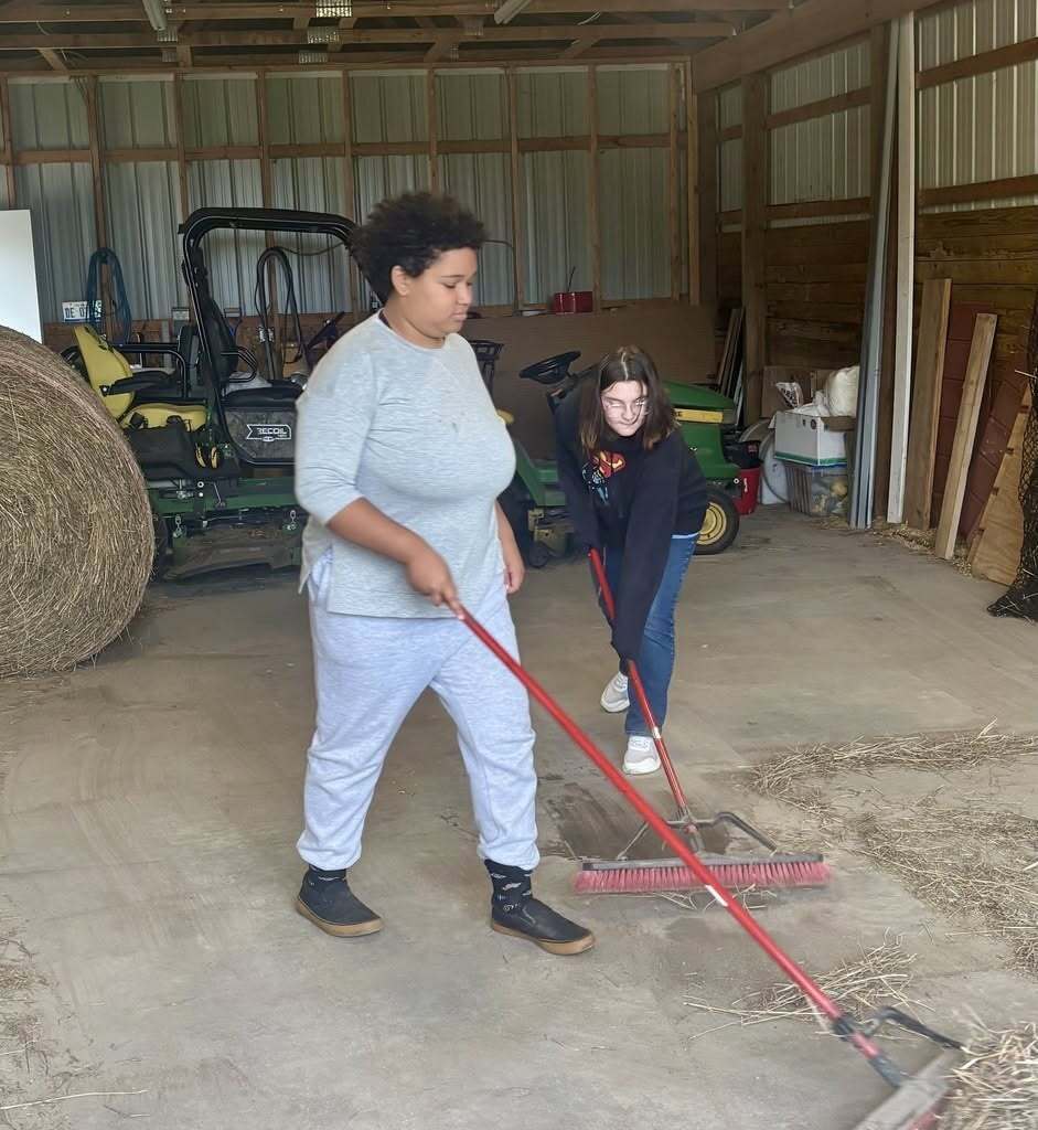 Two individuals are cleaning a floor in a barn. One person is using a broom.