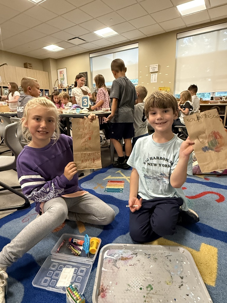 Two children sit on a rug, one holds a paper bag. Nearby, they have open boxes of art supplies. Behind them, children sit at tables in a room.