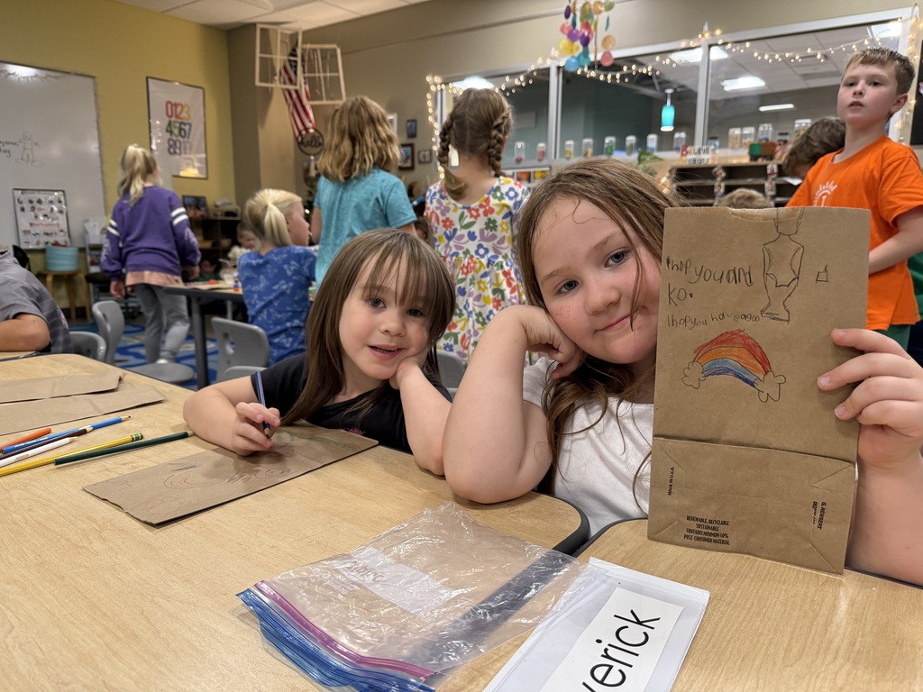 Two girls in a classroom with brown paper bags. One holds a drawing, the other shows a plastic bag with the word "erick."