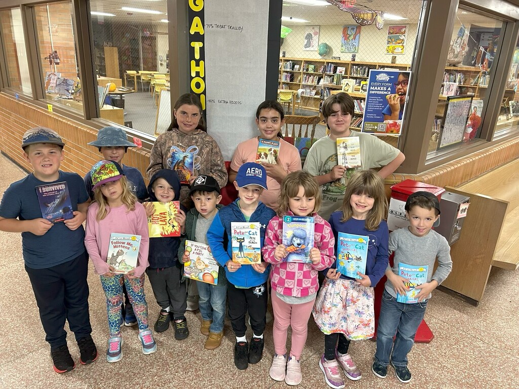 Group of children holding books, standing in a library. A glass wall and bookshelves are in the background.