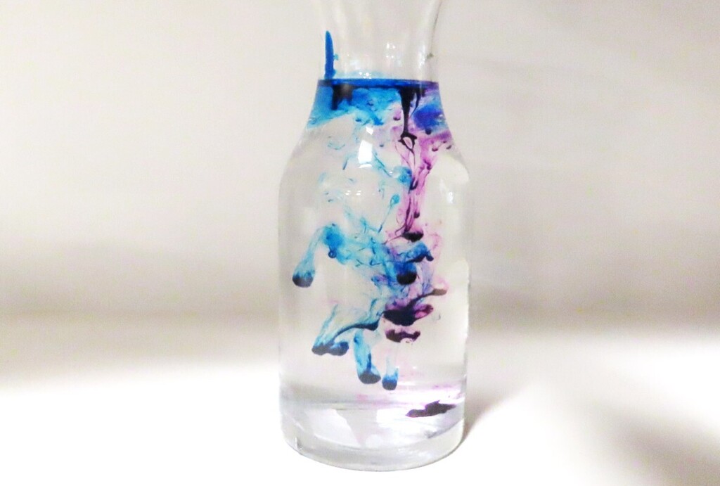 A clear vase with blue, purple, and pink liquid swirling inside, displayed against a white background.