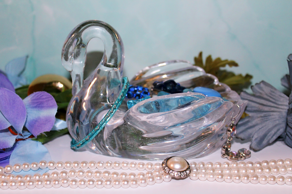 A glass swan figurine, adorned with colorful beads and jewels, rests on a table with flowers and a pearl necklace.