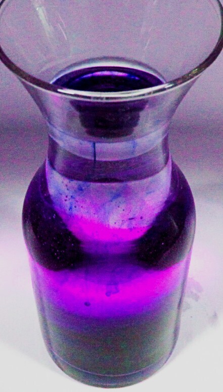 A glass vase filled with purple liquid is displayed on a white background. The vase has a cylindrical body with a wide opening.