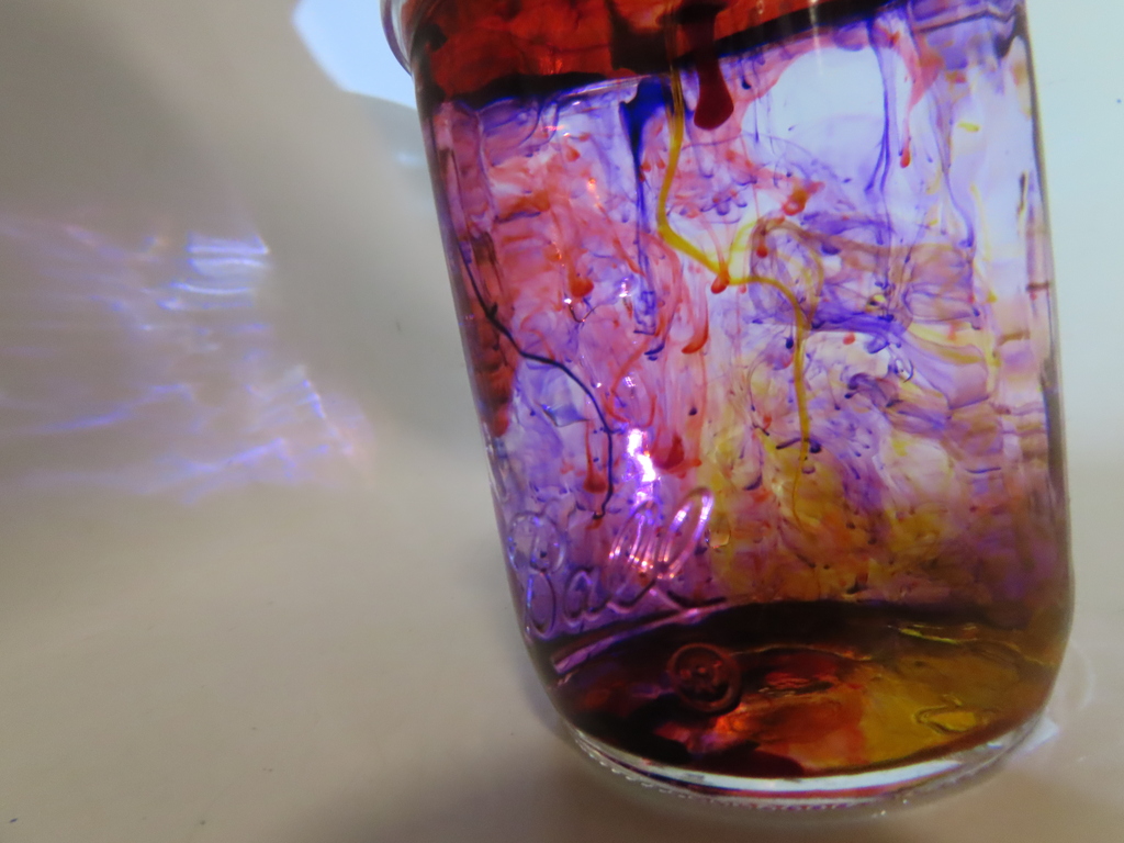 A clear glass jar filled with layers of red, purple, yellow, and blue liquid on a white surface.
