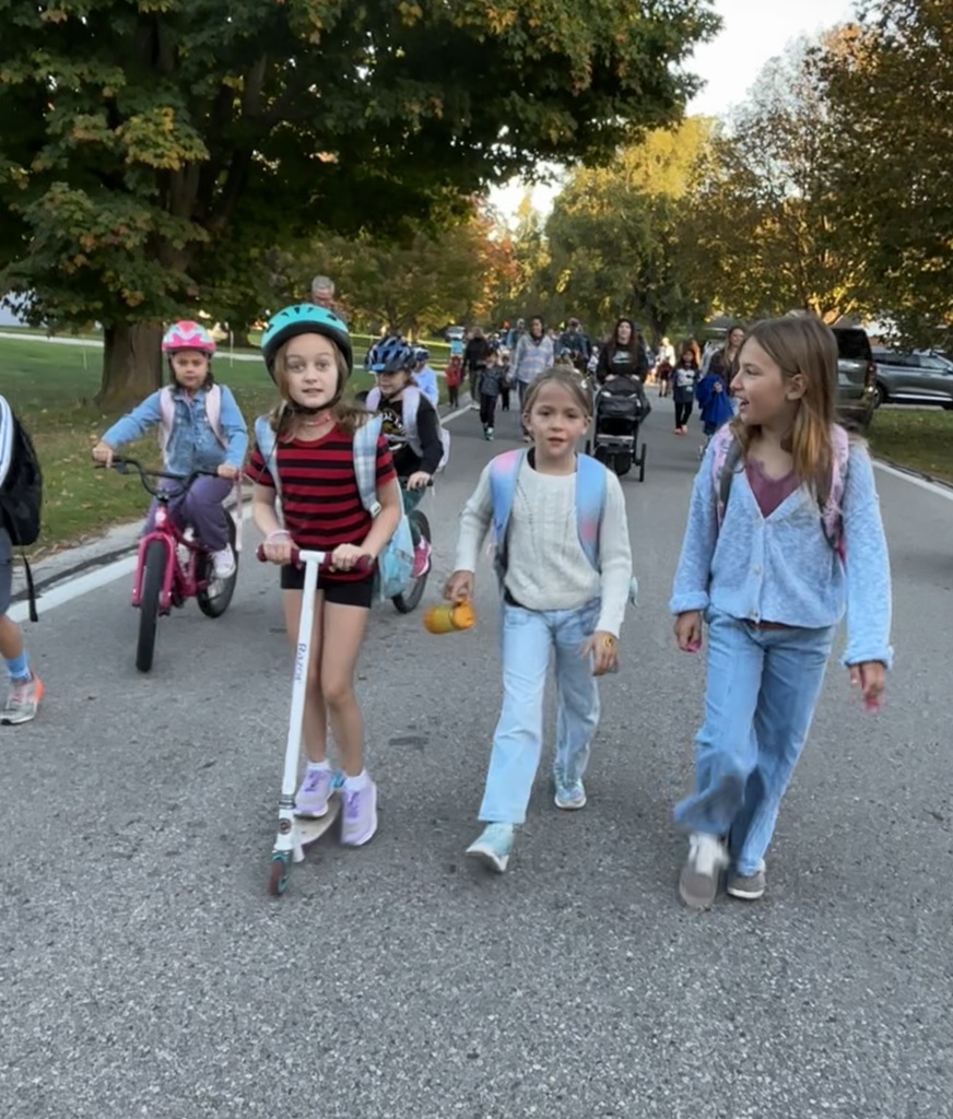 Children walk down a road with green trees in the background. One child rides a scooter, and another rides a bicycle.
