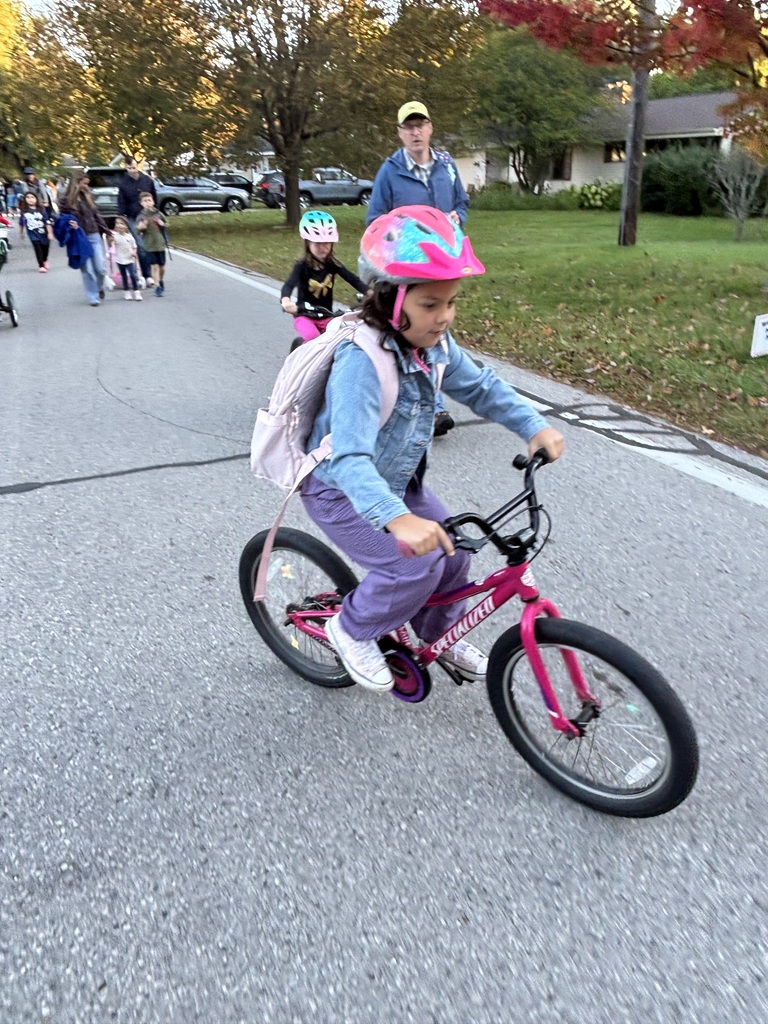 A child in a pink helmet rides a bike down a street, with a person walking behind.