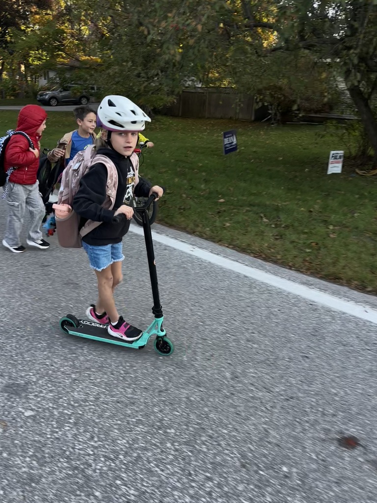 Child on scooter with backpack. Two other children walk behind on sidewalk. Green grass, trees, and signs in background.