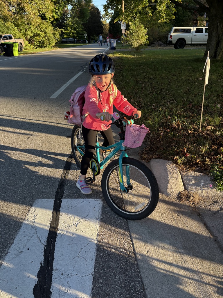 A child in a pink jacket and helmet rides a blue bicycle with a pink basket at a crosswalk.