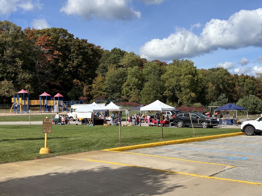 A park with a playground, several tents, and vehicles parked. Green grass, trees, and blue sky with clouds.