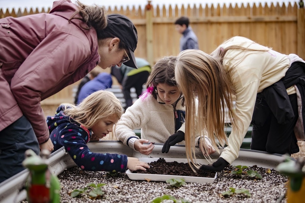 Several children and adults wearing gloves, working with soil in a container in an outdoor garden.