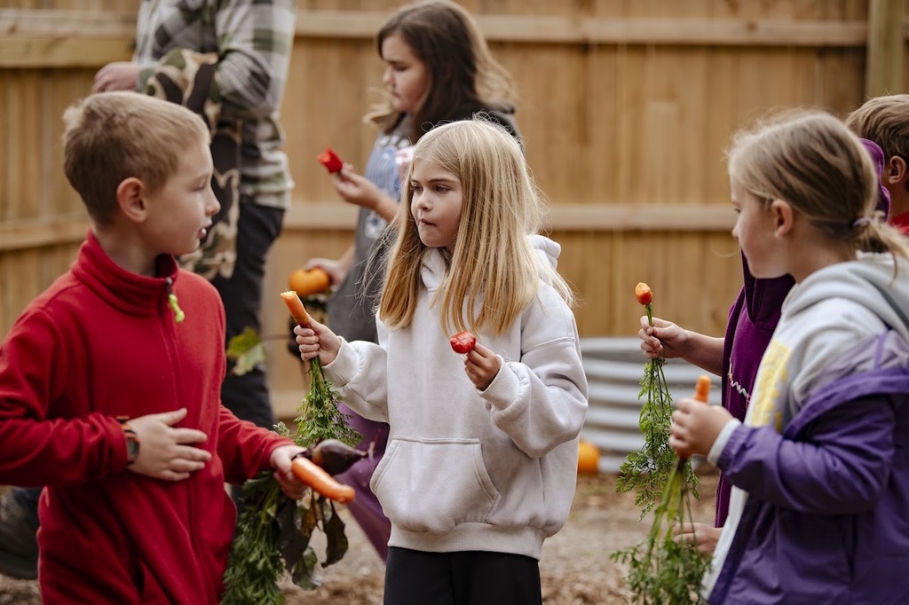 Kids holding carrots stand in a garden. One child in a white hoodie looks at a boy in red.