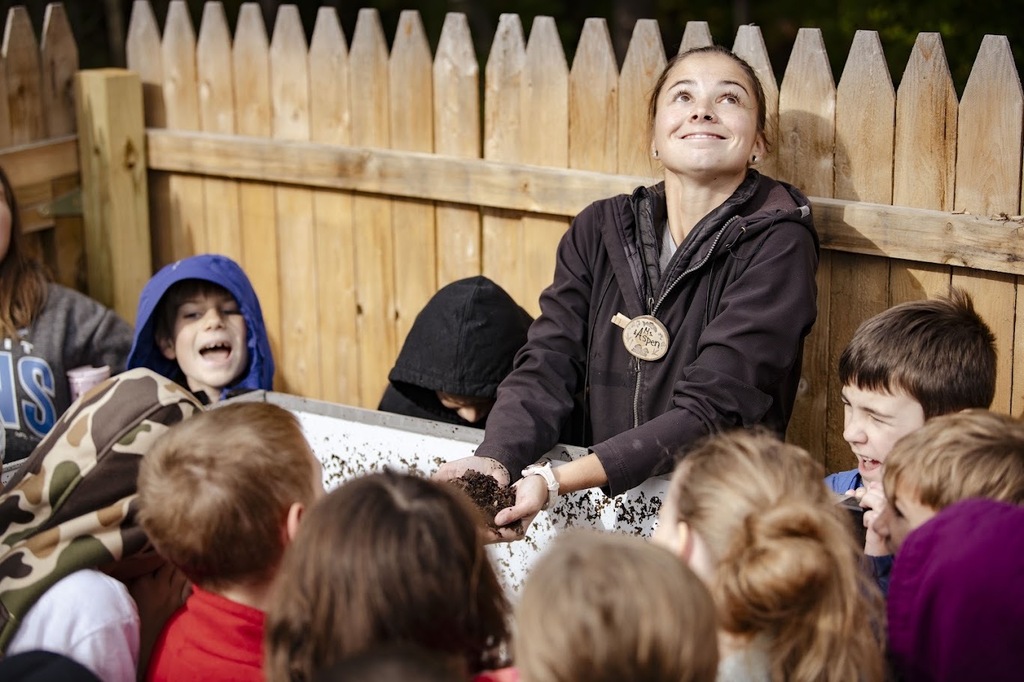 A woman with a medal helps a group of children explore a wooden box. Background: Wooden fence.
