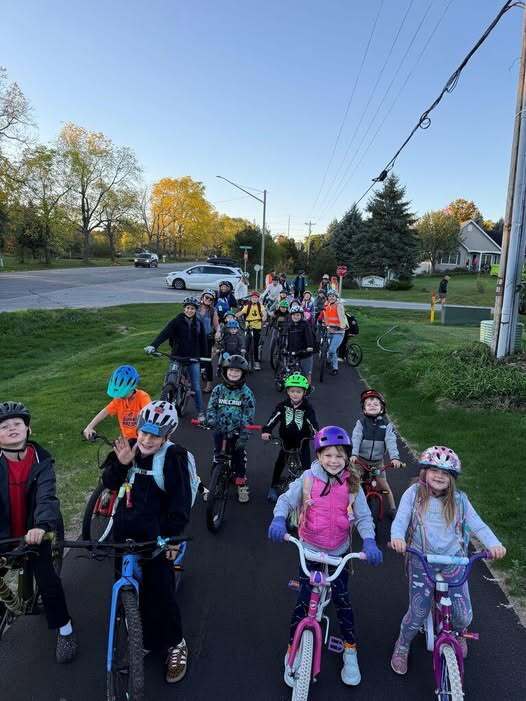 A group of children, wearing helmets, ride bicycles down a paved road. Background shows houses, trees, and electric poles.