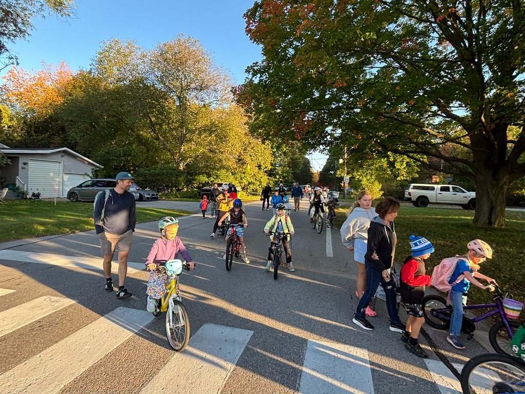 Group of people in a suburban street with trees and houses in the background. Some are riding bicycles and others are walking.