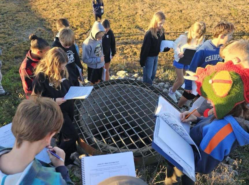 A group of children stands around a metal grate, some holding papers and others looking at it.