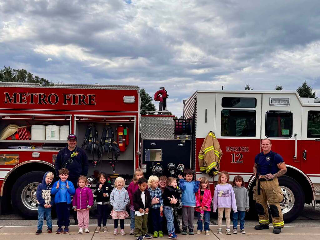 Children stand in front of a red fire truck with a firefighter on the ladder. The sky is cloudy.
