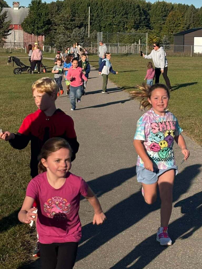 Children run on a grassy path; one leads, others follow. Shadows fall on the ground. Trees and buildings in the background.