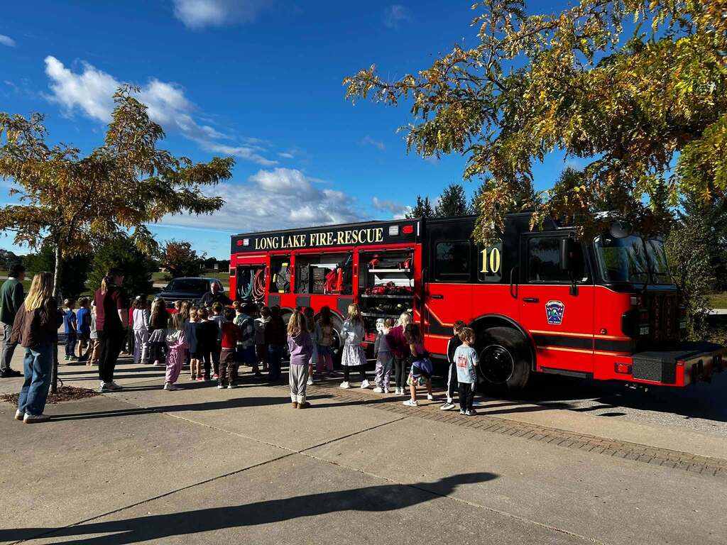 A red fire truck labeled "Lake Fire Rescue 10" is parked. People, including children, gather around it. Trees with leaves are in the background.