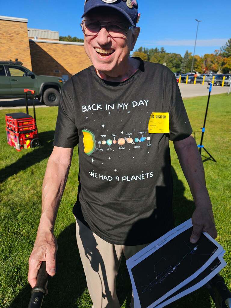 A man in a cap holds a paper and smiles at the camera. He wears a shirt saying "Back in my day, we had 9 planets."