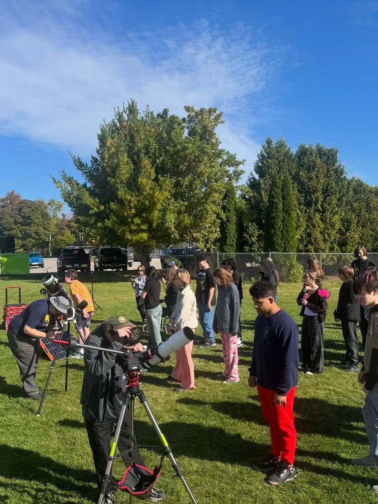 A group of people are gathered on a grassy field with telescopes, observing the sky on a sunny day.