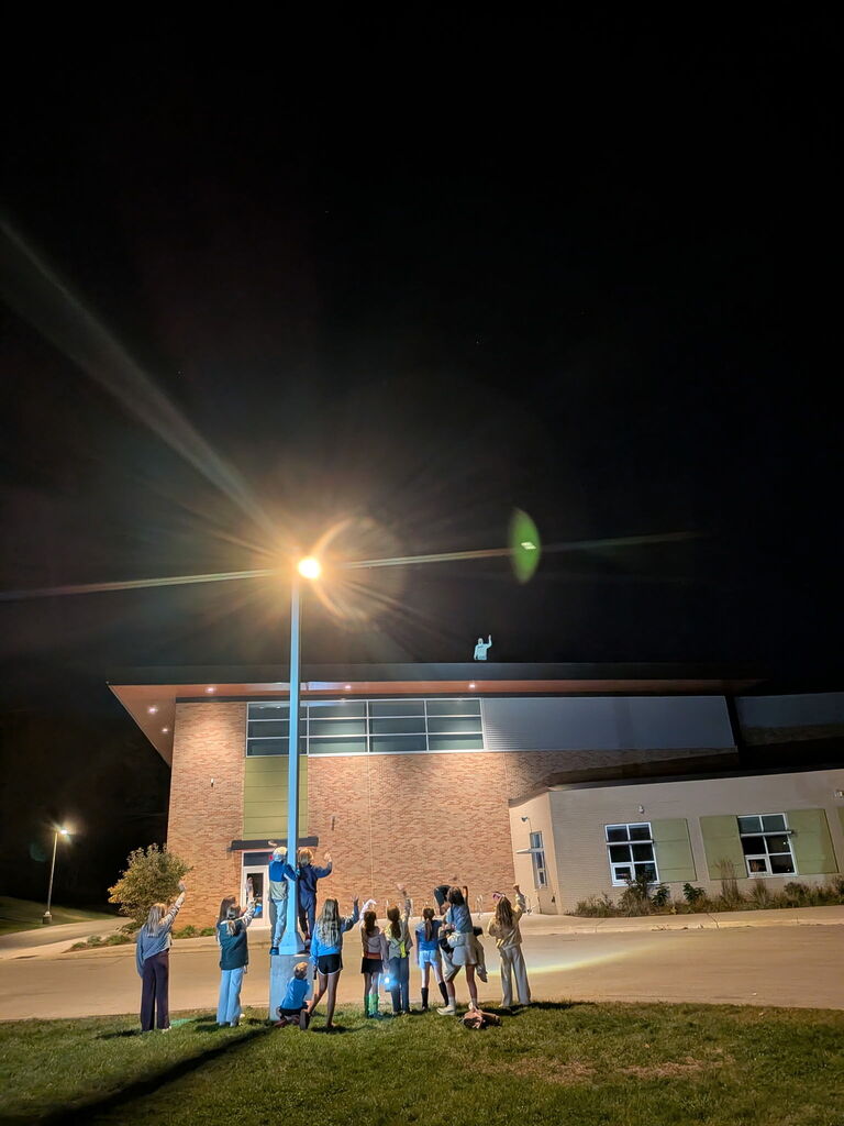 Group of people waving up to Mr. Swelges on the roof in front of the school at night..