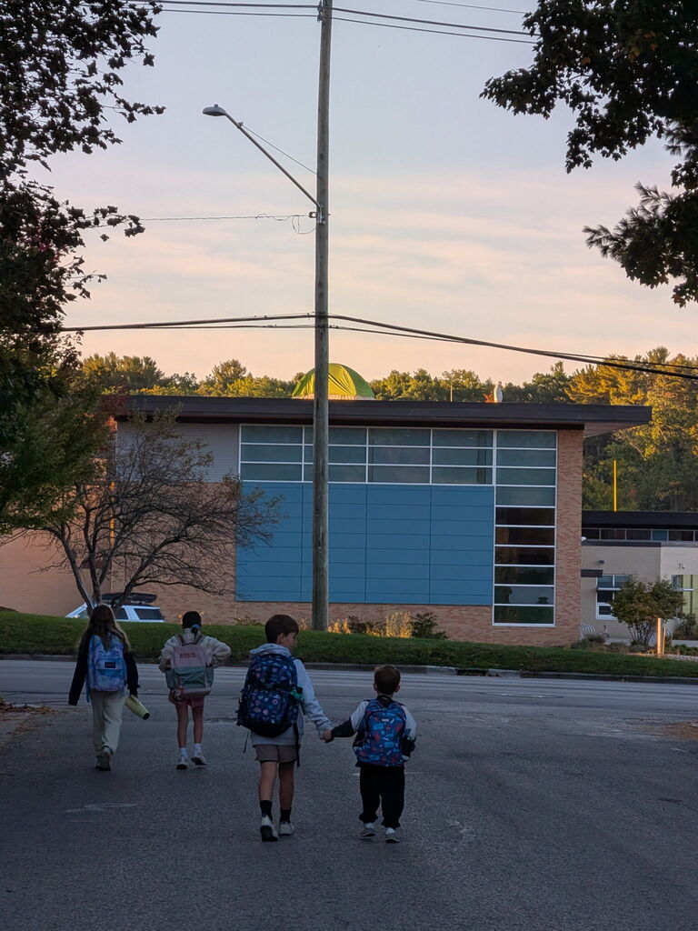 Four children with backpacks walk across a street. Behind them, the school building has a tent on top of the building