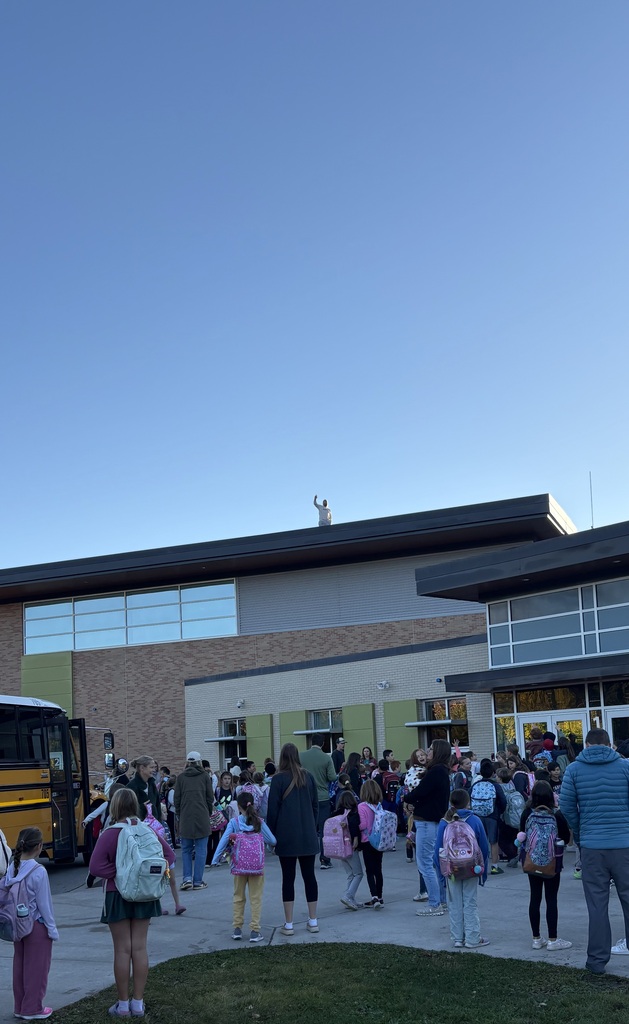 A group of students wearing backpacks stand near a school building with large windows. A bus is parked and crowd is waving to man on top of the building