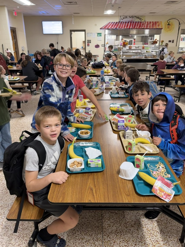 Children seated at a cafeteria table with trays of food. Some children have backpacks.
