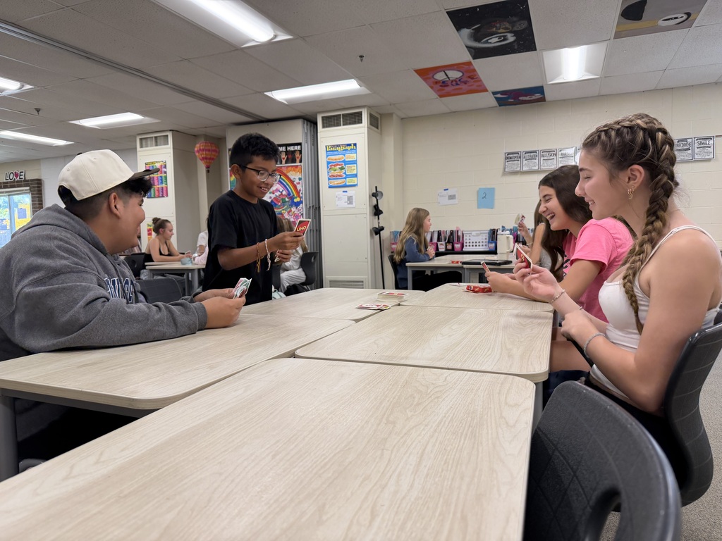 Students in a classroom. Three are seated at a desk, engaged in conversation, and another is standing.