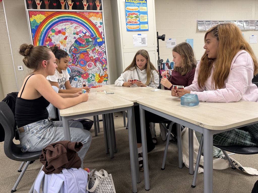 Five women sit at tables in a classroom. One woman holds a glass. Colorful posters are on the wall behind them.