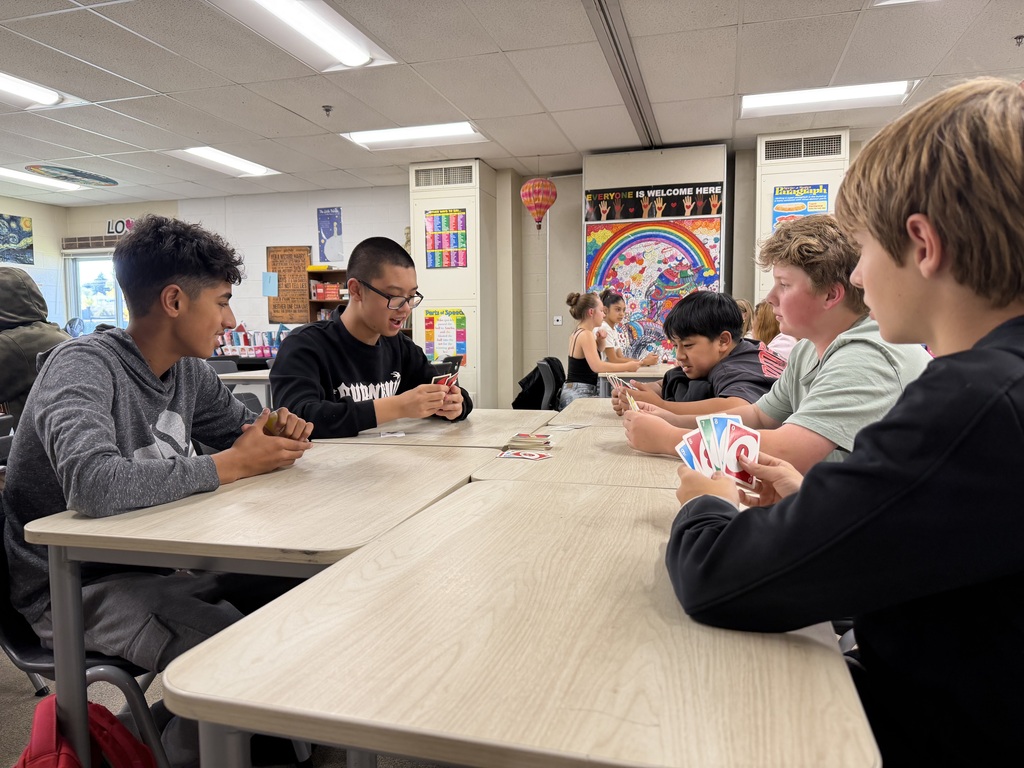 Six students seated around a table in a classroom. Each holds playing cards and appears to be engaged in a card game.