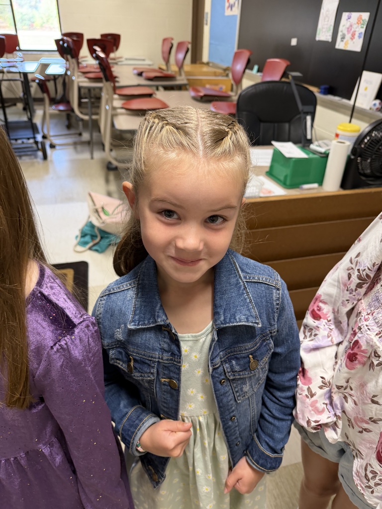 A young girl with blonde hair and a denim jacket smiles in a classroom with desks.
