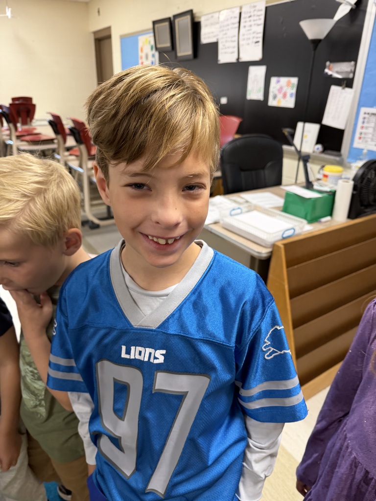 A boy in a blue Lions football jersey smiles and stands with other children in a classroom.