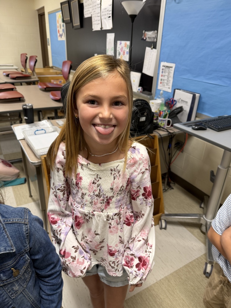 A girl with blonde hair smiles in a classroom. Behind her are desks, a blue wall, and various classroom items.
