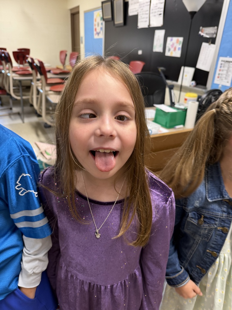 A young girl in a purple dress with her tongue out stands between two others in a classroom.
