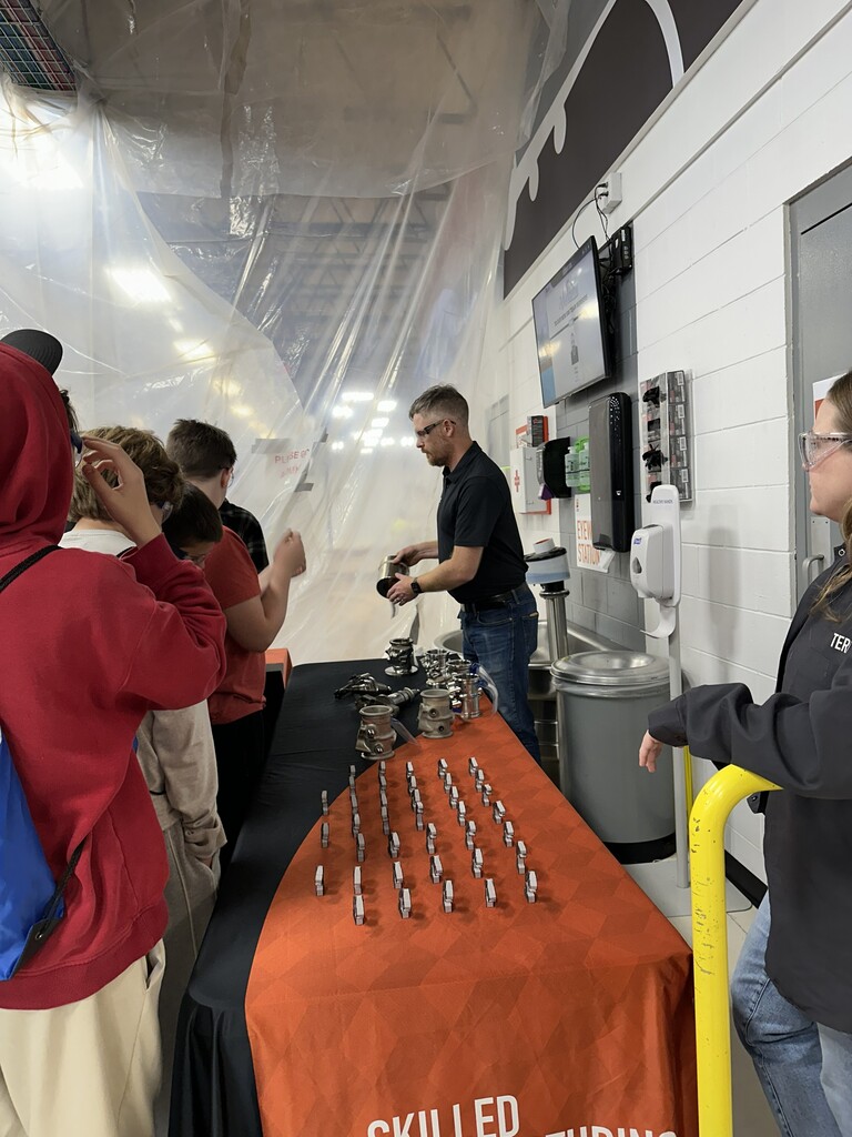 A group of people stands around a table displaying metal objects. One man uses a machine.