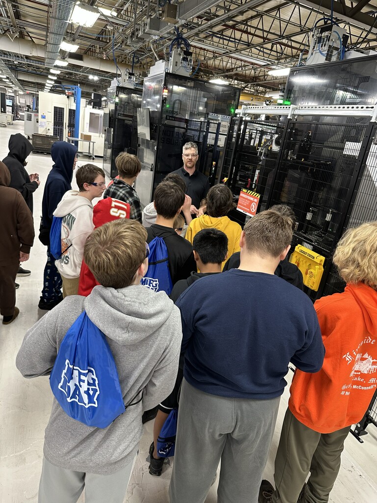 A group of people, including children, are standing in an indoor factory setting, watching a person speaking.