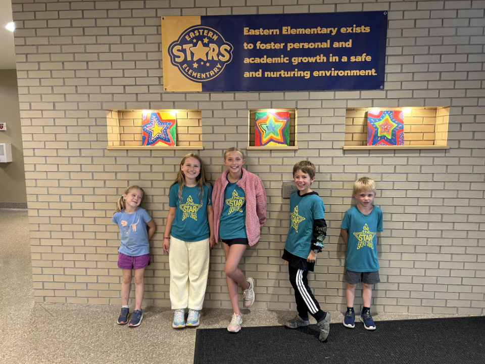 Five children stand in front of a wall with a blue sign. The children wear matching teal shirts. Three colorful star designs are displayed above.