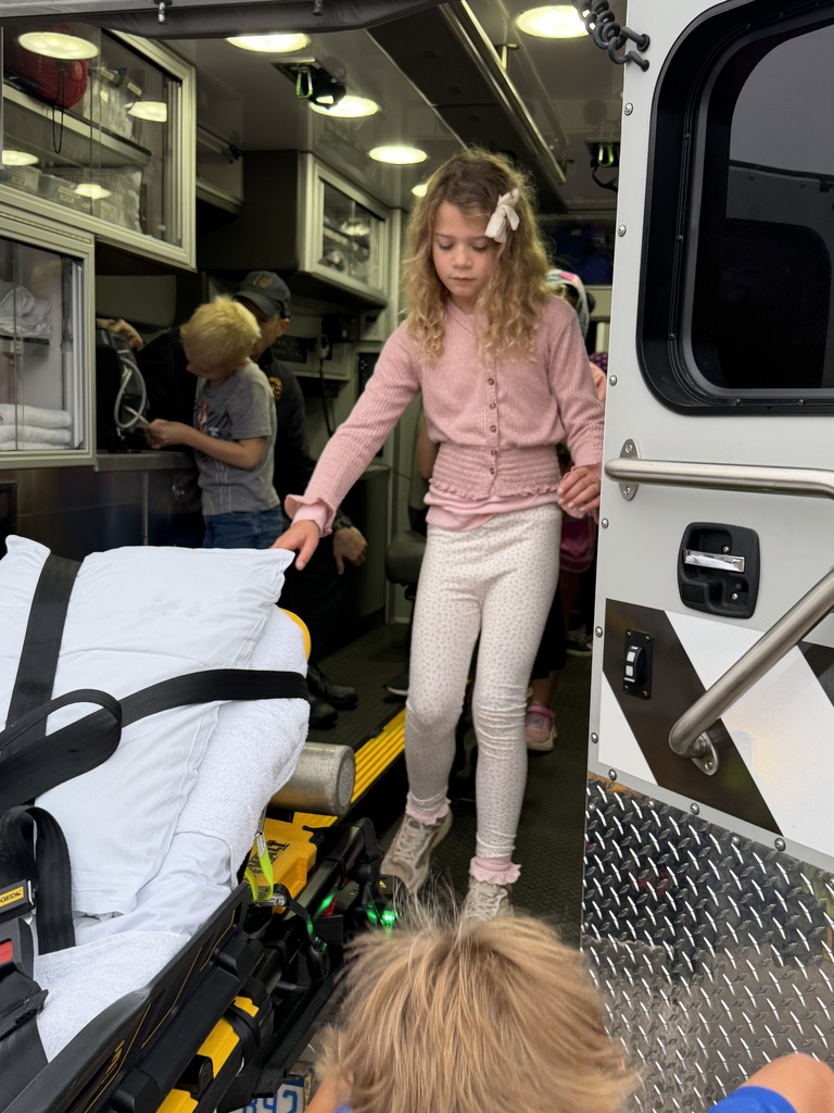 A child in pink holds onto an ambulance handrail as another child sits on the stretcher.