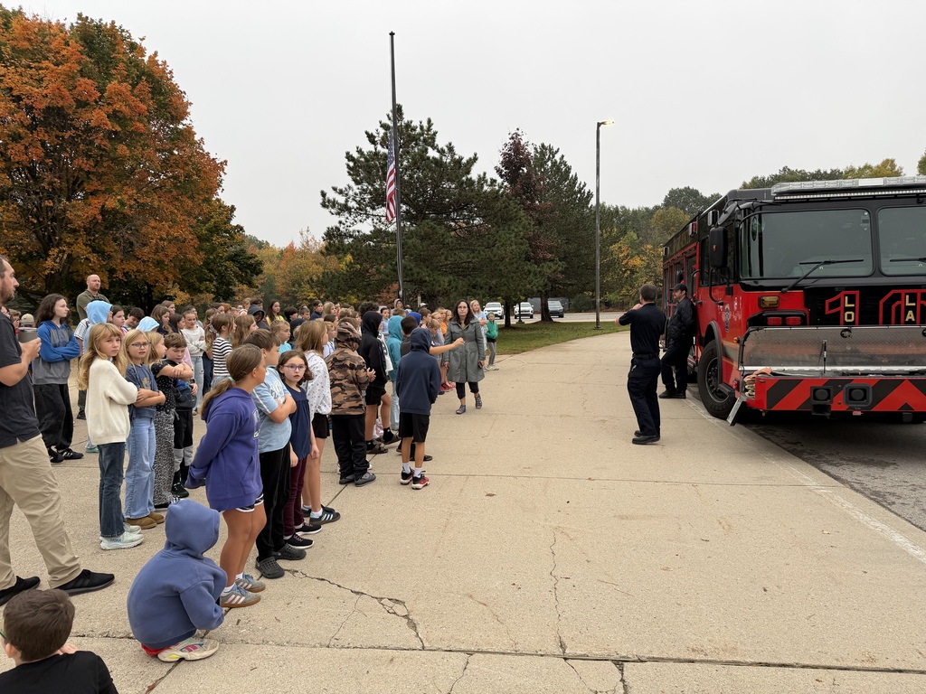 People stand along a sidewalk with a firetruck parked on the side, a person speaks to the crowd.